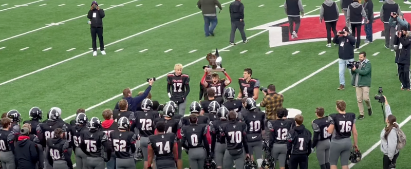 C-FC football team huddled and raising silver ball trophy at Camp Randall